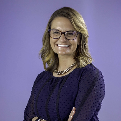 Portrait of Kate Kavan smiling with crossed arms against a light-purple background.