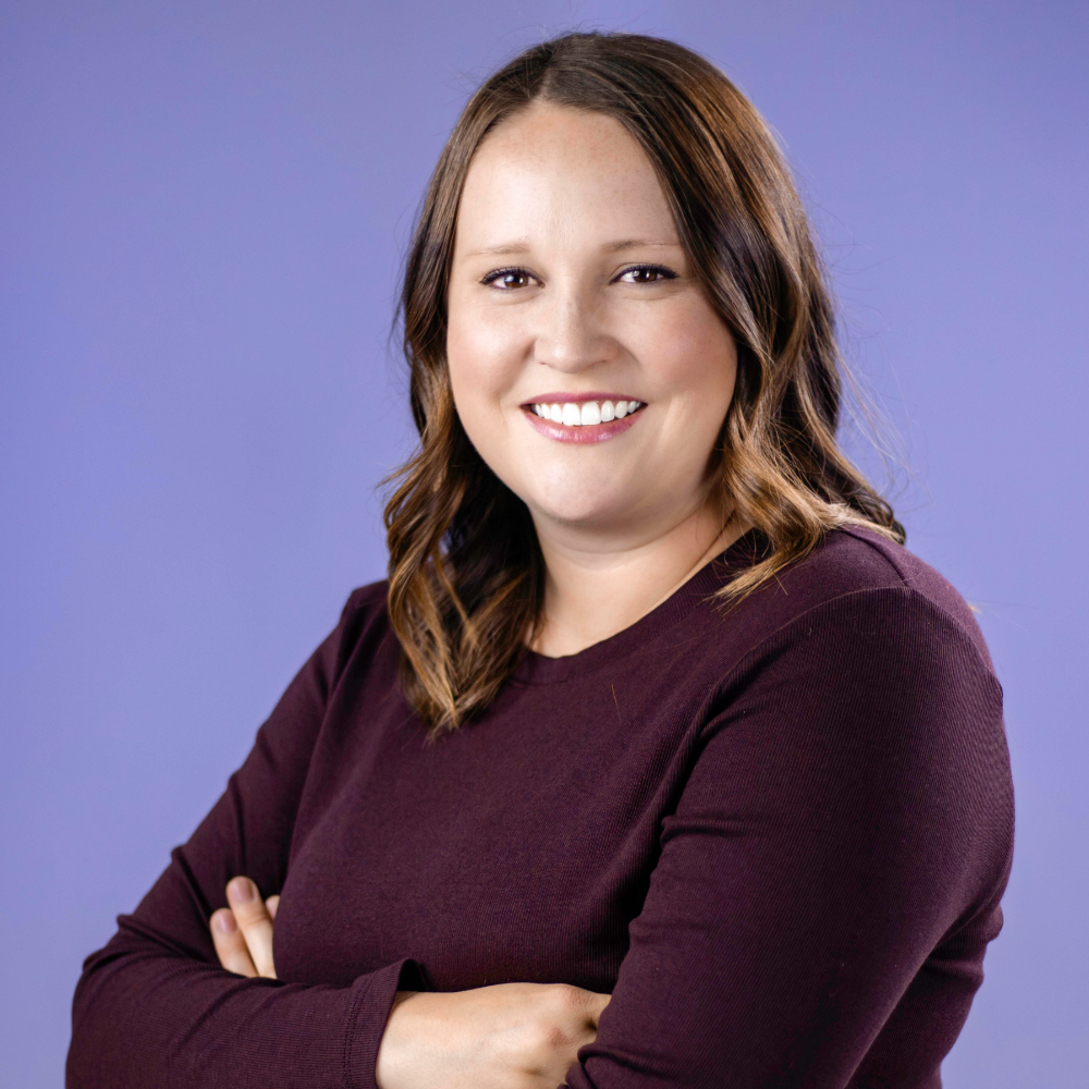 Portrait of a smiling Lauren Baak with brown, shoulder-length hair against a purple background. She is wearing a dark plum-colored top and have their arms crossed.