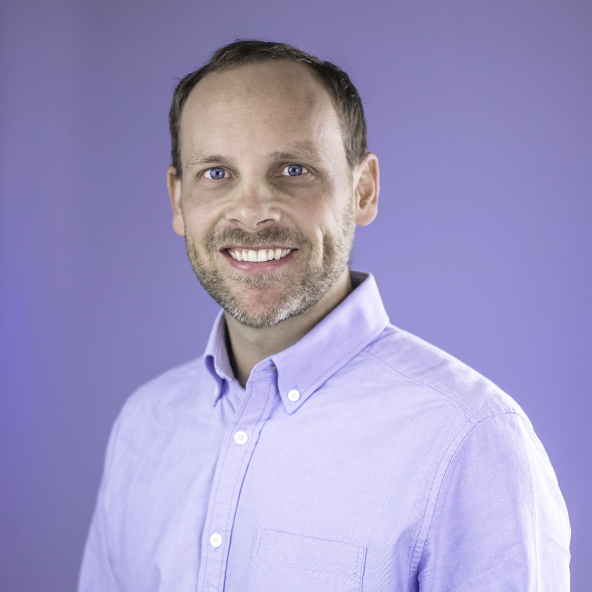 Portrait of a smiling William Mauldin in a light blue shirt against a light purple background.